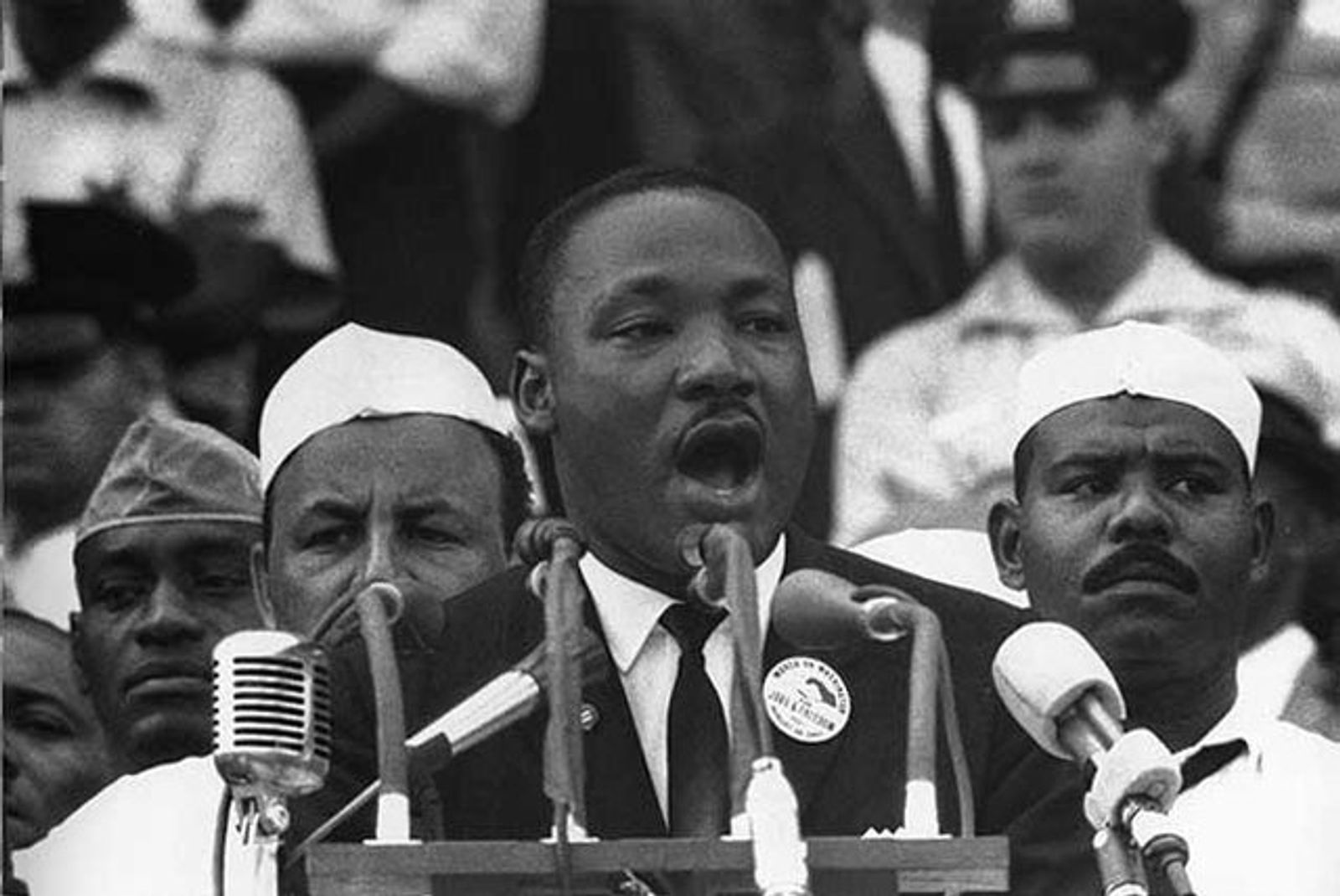Black-and-white photograph of a man speaking intently into a bank of microphones at a rally, flanked by men in white caps.