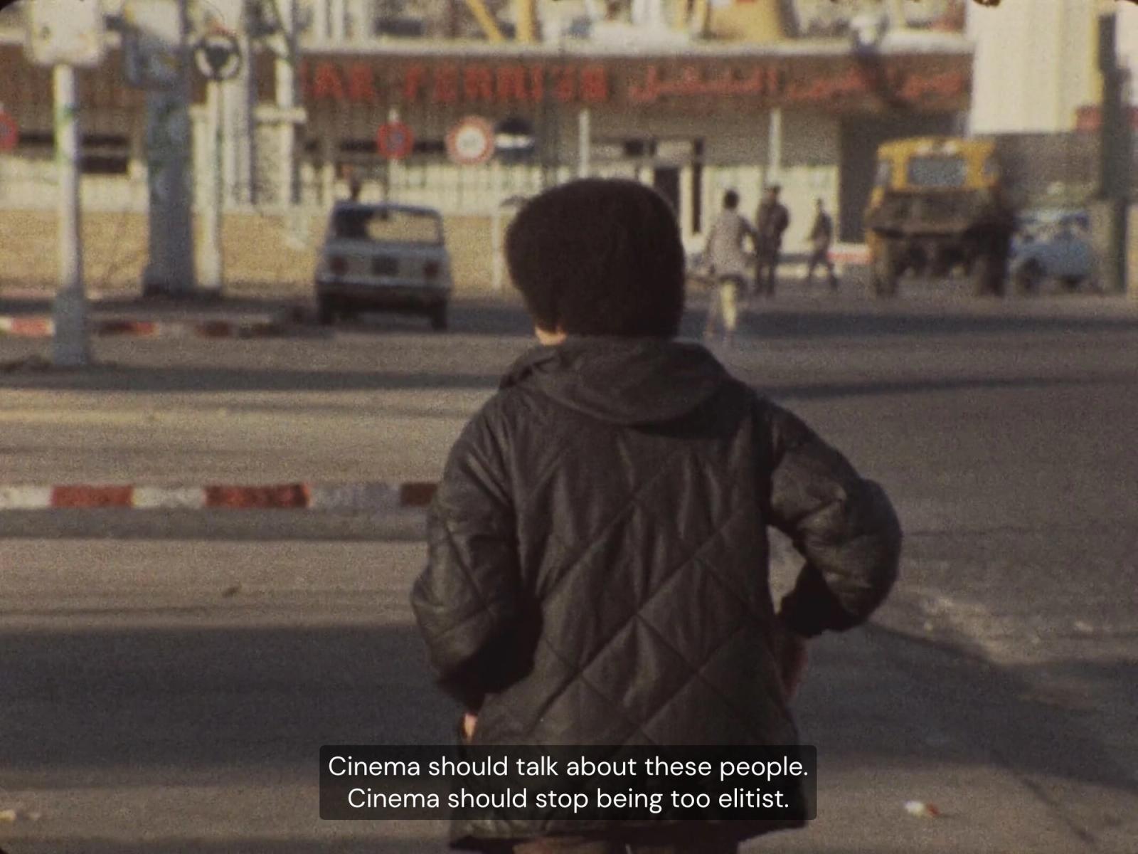 Film still of a child in a quilted jacket seen from behind in a street, with subtitles calling for cinema to stop being elitist.
