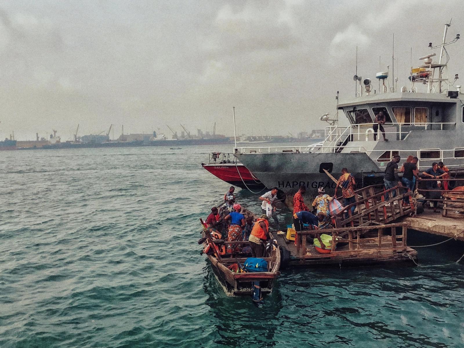 People move from a small wooden canoe onto a jetty beside a larger boat on grey-green water in Lagos.