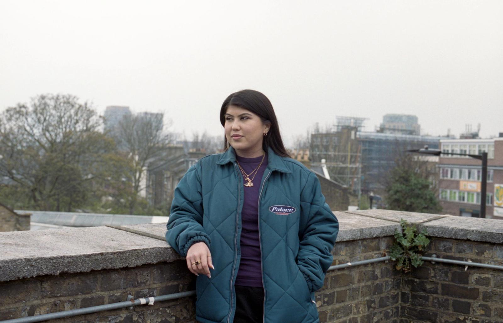 Freshta, in a teal quilted jacket, leans on a rooftop wall and looks off to one side, with a city skyline behind her.