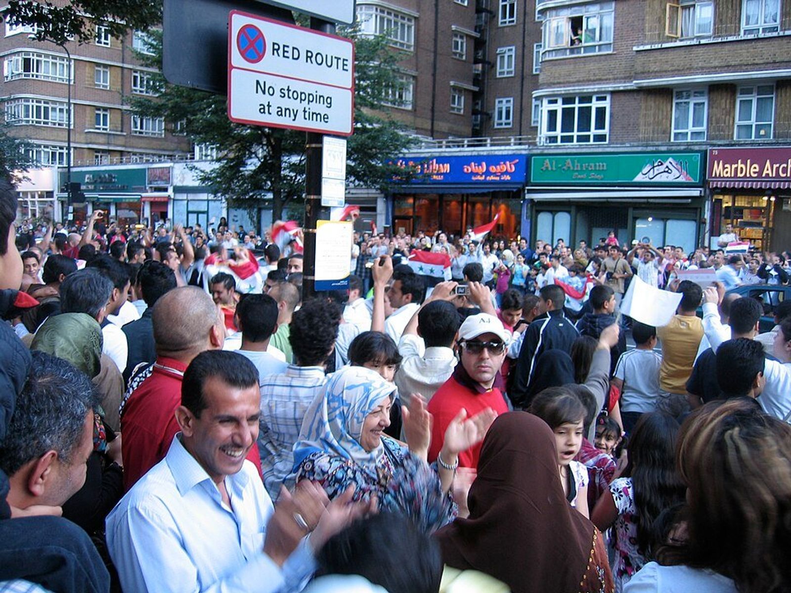 Fans celebrate in a crowded street outside Arabic shopfronts, clapping and waving flags beneath a Red Route sign.