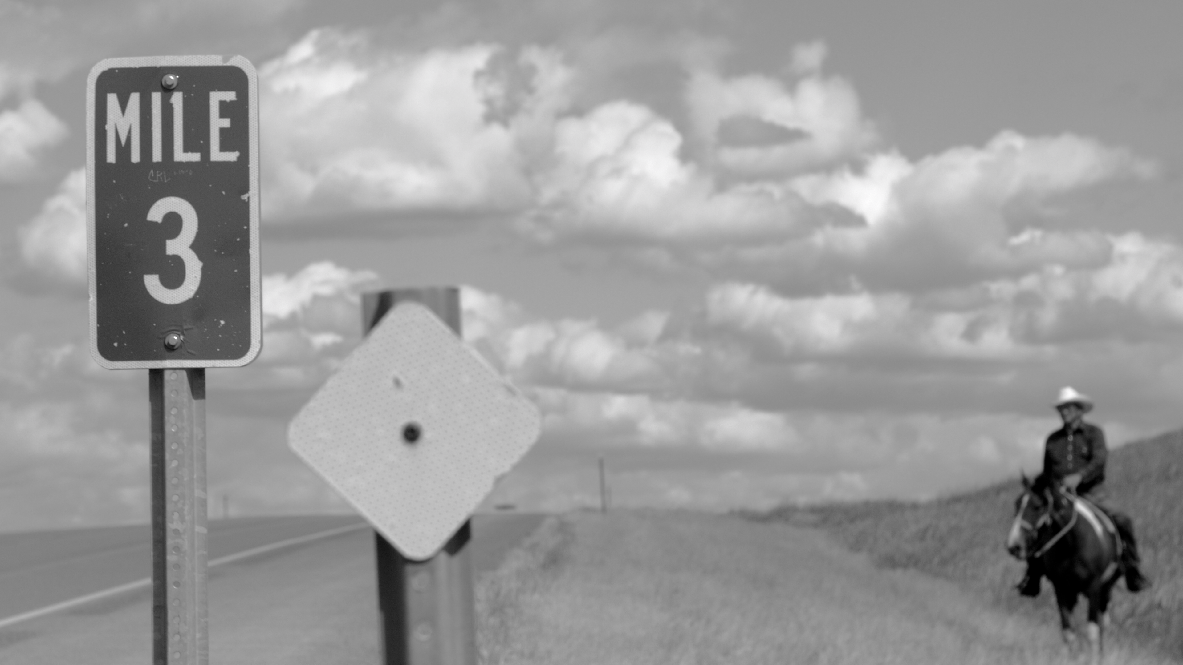A Mile 3 highway sign in focus, a man on horseback out of focus, in the background to its left.