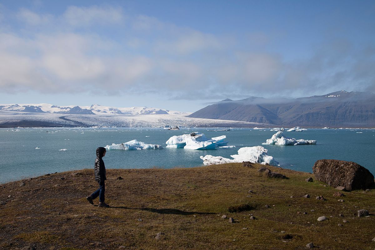 A child walking on a grassy hill in front of a body of water with icebergs and a snowy mountain range