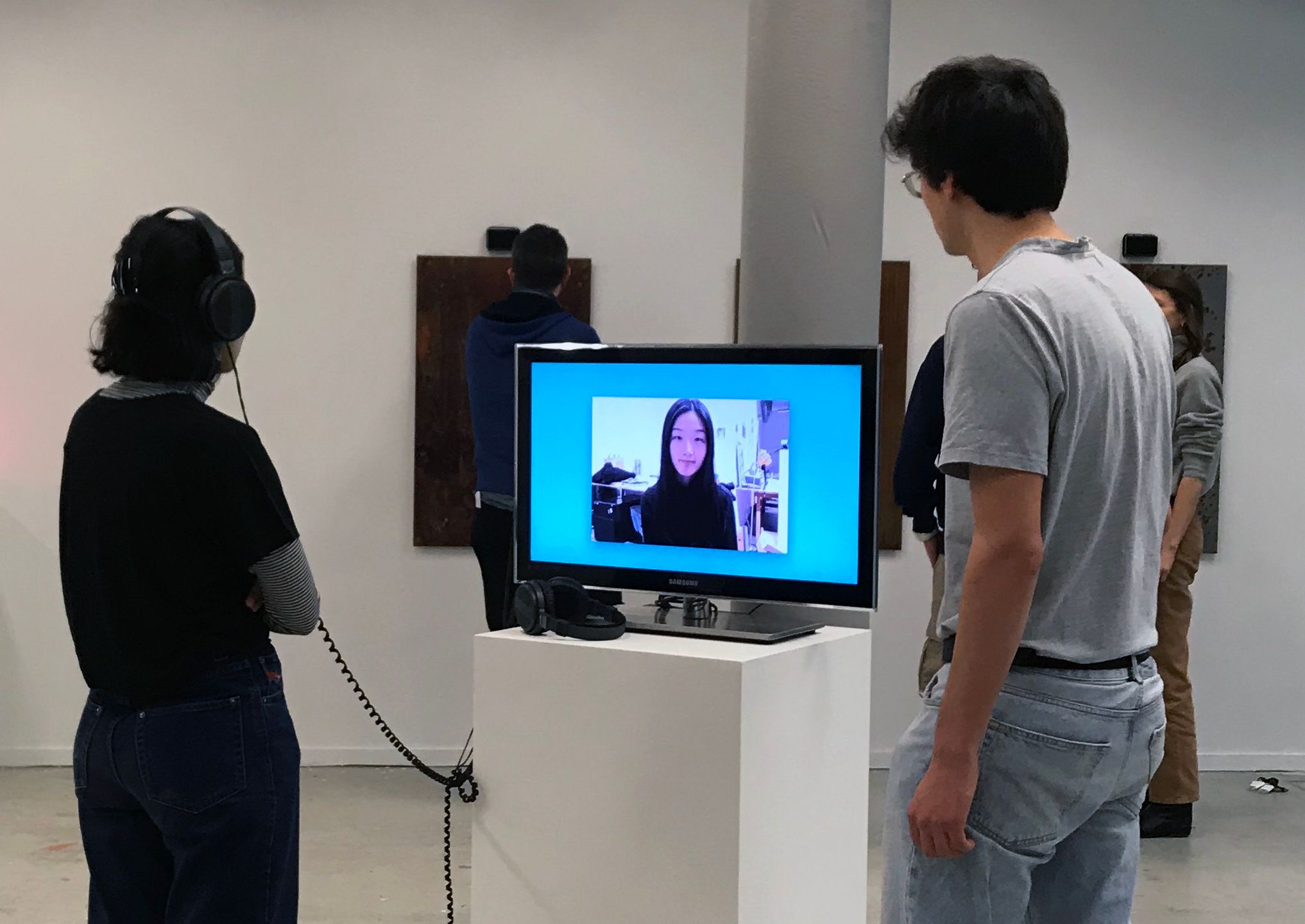 Two people watching cage-shining-pearl on a monitor in a gallery; one listens with headphones