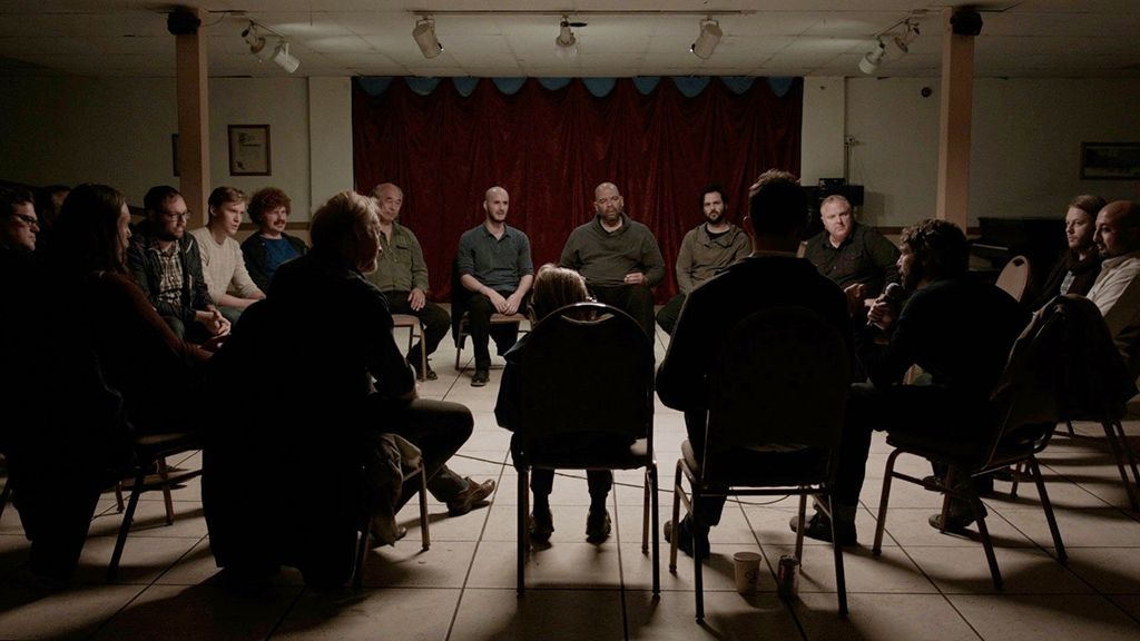 A room of men sitting in folding chairs a circle, support group style