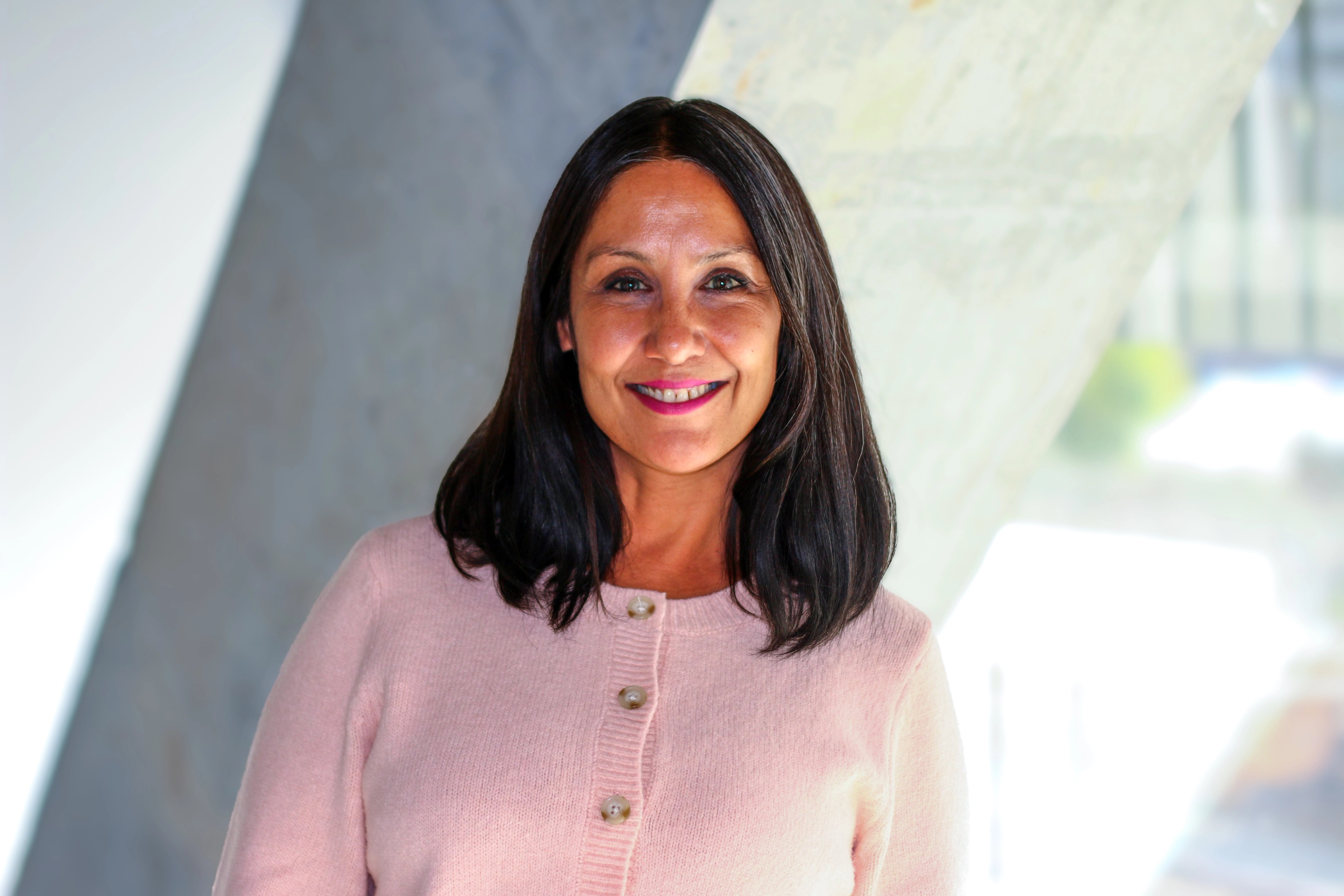 Portrait of Sharon Cheema from Ava Industries smiling, standing in a bright, modern office space wearing a light pink cardigan.