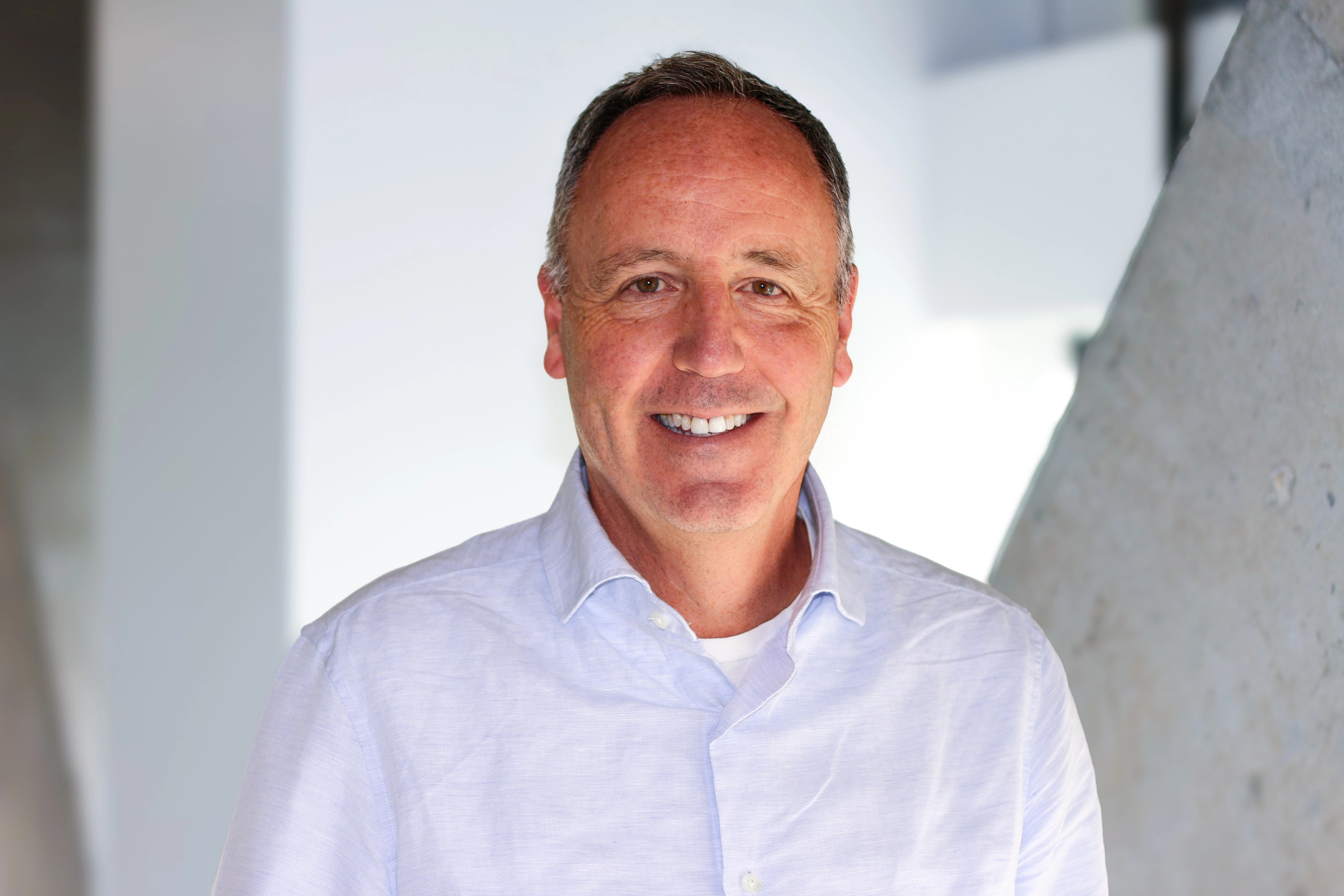 Portrait of Rob Bevis from Ava Industries, smiling in a bright office setting, wearing a light shirt and standing in front of a modern angled concrete wall.