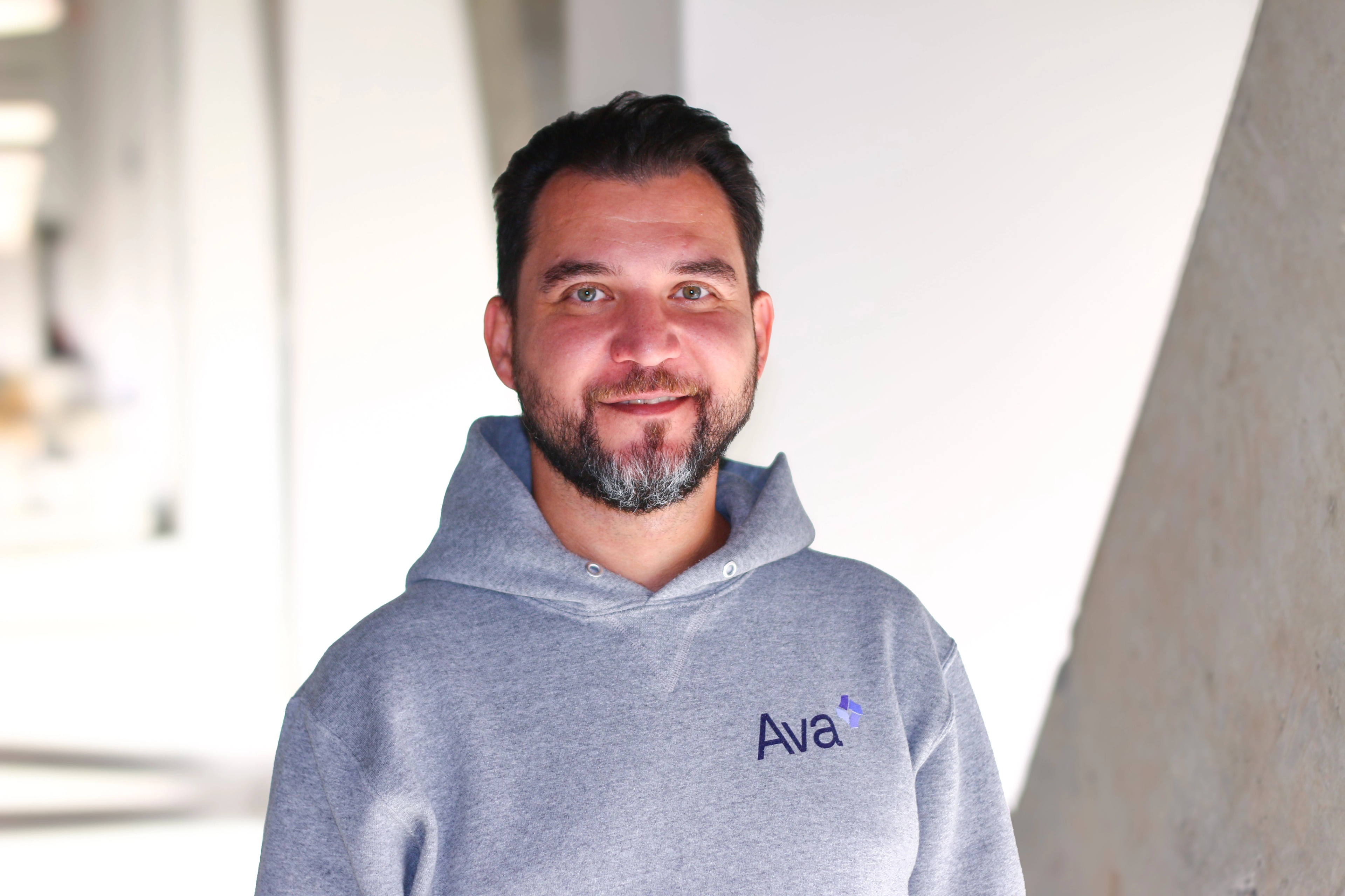 Portrait of Raed Tulefat, Software Developer at Ava Industries, smiling while wearing a grey hoodie with the “Ava” logo, standing in a modern office space with concrete architecture in the background.