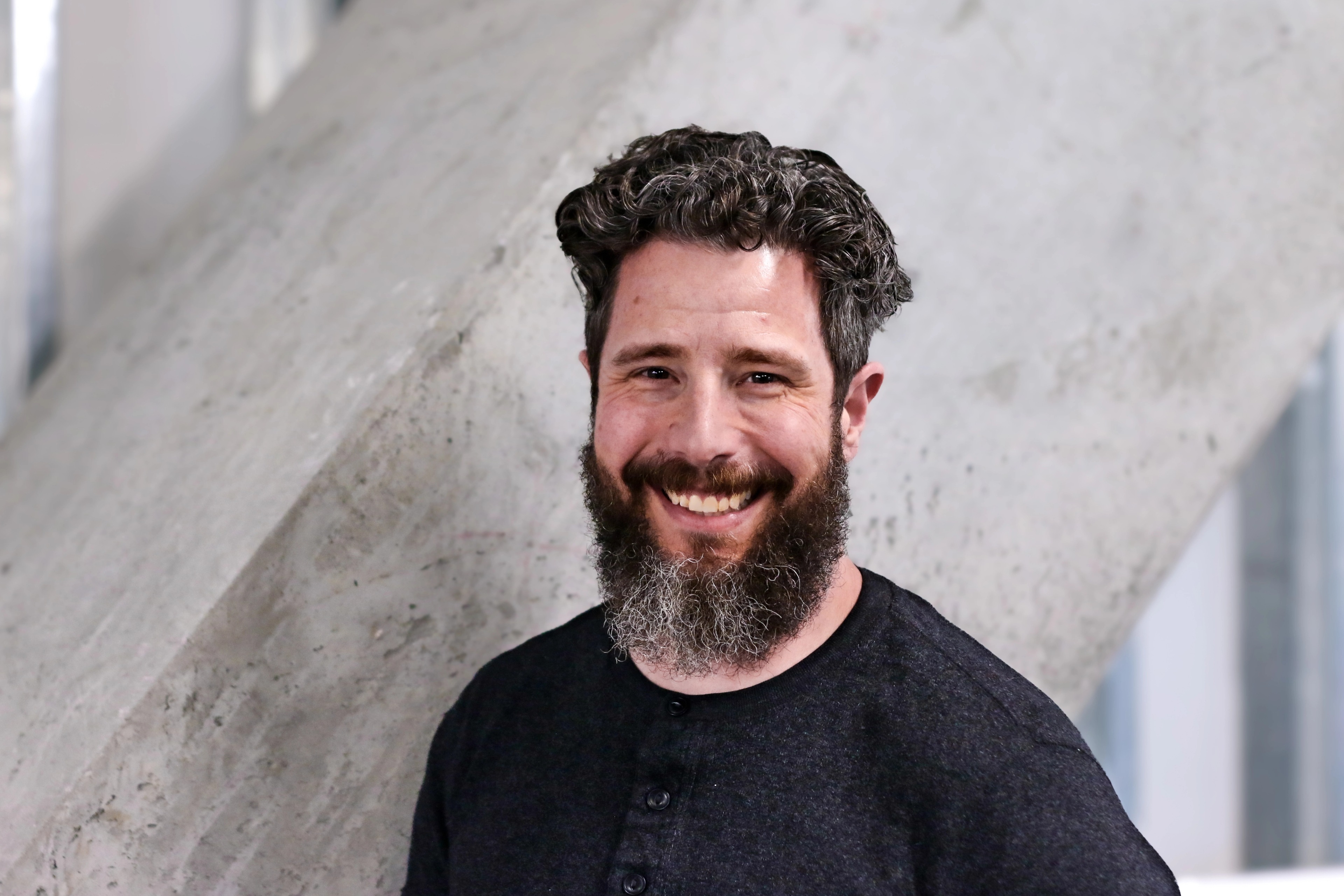 Portrait of Will Forseth, Accounting Analyst at Ava Industries, smiling, with a full beard and medium-length curly hair, wearing a dark henley shirt, standing in front of a light modern office background.