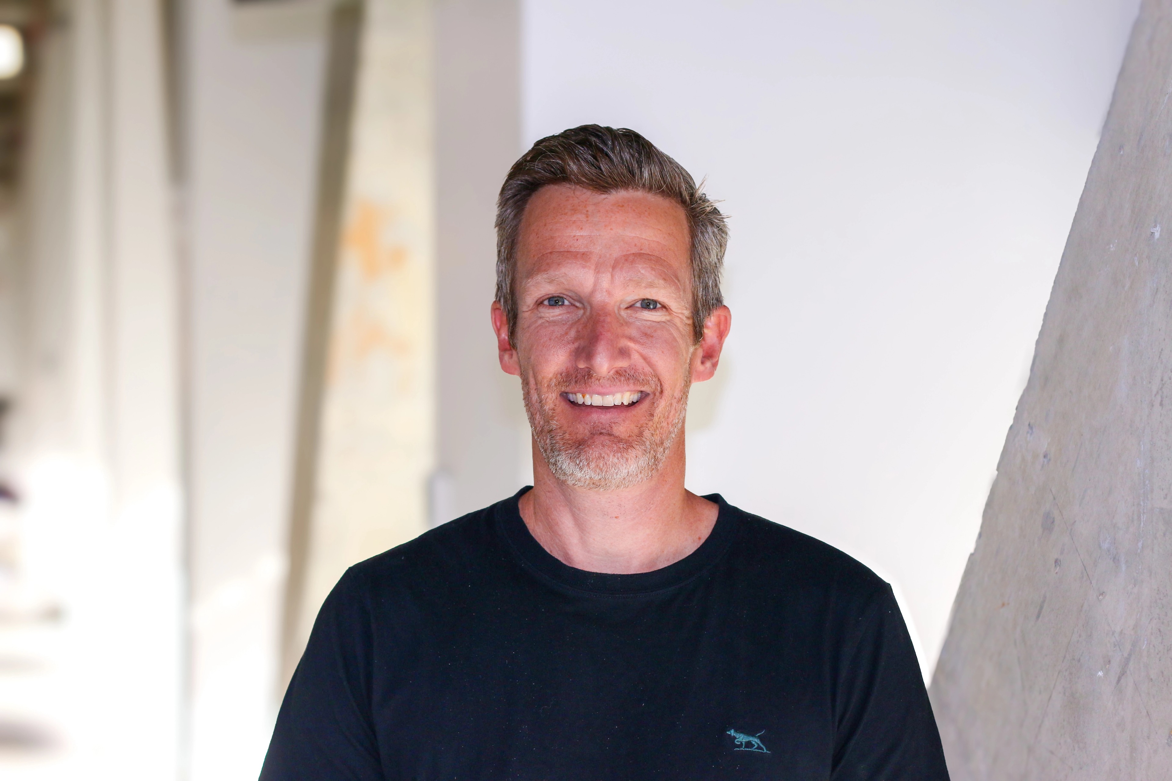 Portrait of Dr Mike Forseth from Ava Industries, smiling in a modern office environment, wearing a dark shirt and standing in front of a light-coloured background with soft concrete architecture.