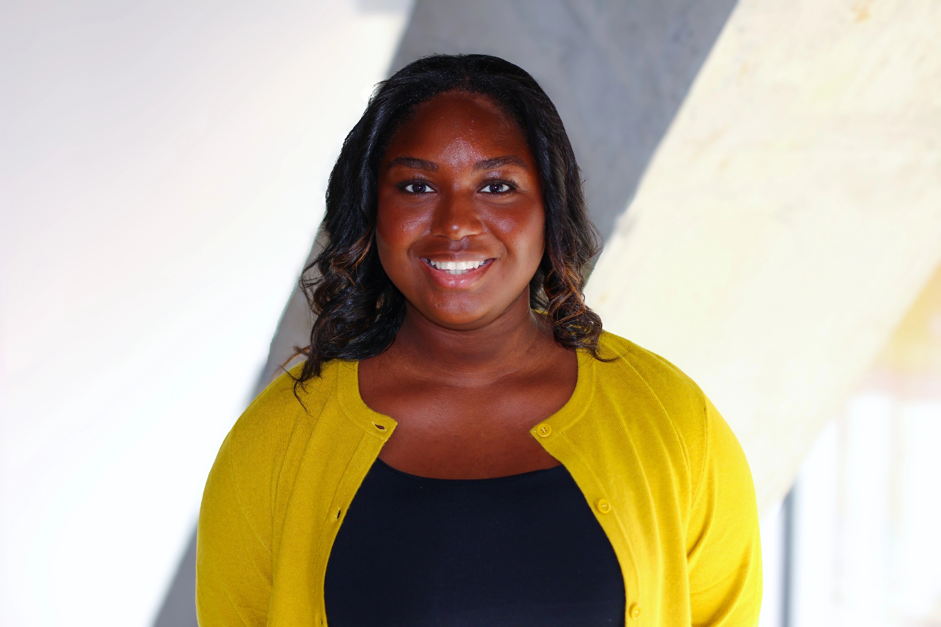 Portrait of Mariam Ukaria from Ava Industries smiling, wearing a yellow cardigan over a black top, standing in a bright modern office environment.