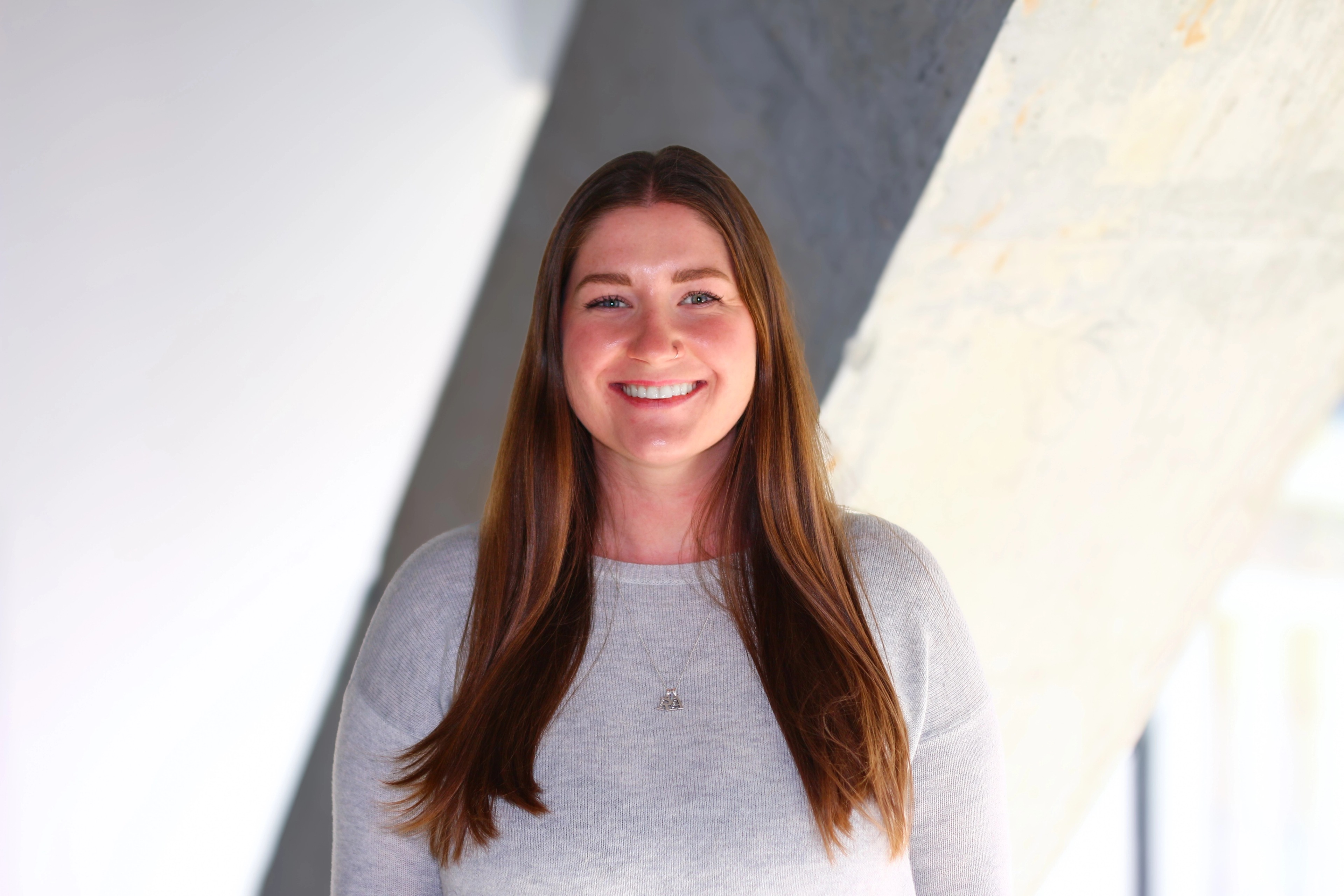 Portrait of Hannah Morin from Ava Industries, smiling in a modern office setting with long brown hair, wearing a light grey sweater.