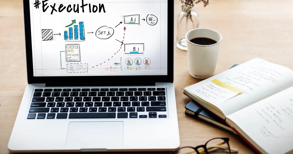 A laptop displaying a project execution plan with flowcharts and diagrams, accompanied by a notebook, a cup of coffee, and glasses on a wooden desk.