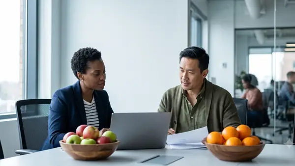 Two diverse executives in smart casual attire review data on a laptop at a conference room table, with a bowl of apples in front of one and a bowl of oranges in front of the other.
