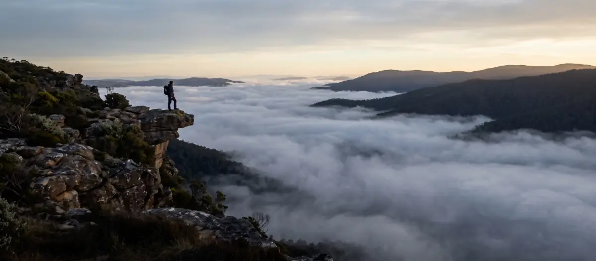 A hiker standing near a cliff edge at dawn, looking out over a fog-filled valley.