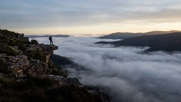 A hiker standing near a cliff edge at dawn, looking out over a fog-filled valley.