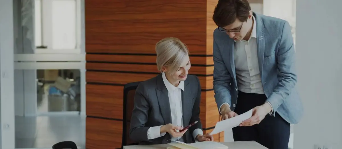 Two businessmen in formal attire sitting at a table, closely reviewing printed documents together in a bright office setting