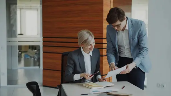 Two businessmen in formal attire sitting at a table, closely reviewing printed documents together in a bright office setting