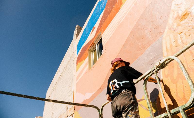 Una muralista con gorra pinta desde una plataforma elevadora la fachada de un edificio en tonos rosas y azules.