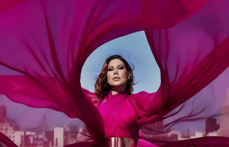Una cantante con vestido fucsia posa entre telas del mismo color que ondean al viento bajo el cielo.