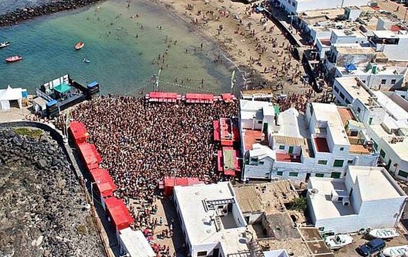 Aerial view of a crowd filling the square by the beach in Caleta de Famara, among white houses.