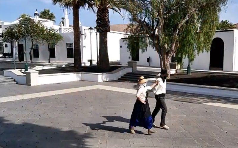 A couple in traditional dress dance in the square in Yaiza, among palm trees and white houses.
