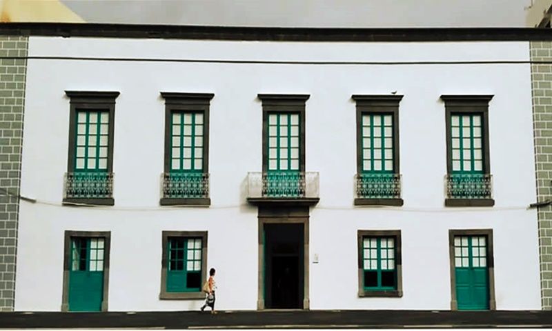 Fachada blanca de dos plantas con ventanas verdes y balcones de forja en Arrecife; una mujer pasa delante.