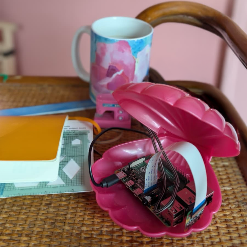 Raspberry pie module in a pink clamshell on a desk in a pink room next to a yellow notebook.