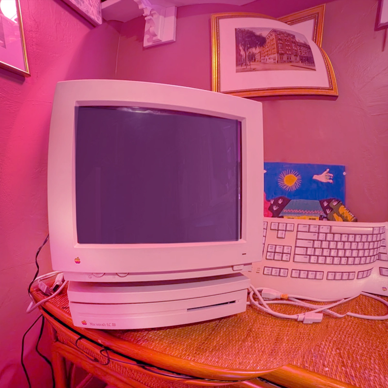 A vintage Apple Macintosh LC III computer and keyboard on a woven desk in a pink room. A framed vintage Troy, New York postcard hangs above, with a colorful child's painting partially visible behind the keyboard.