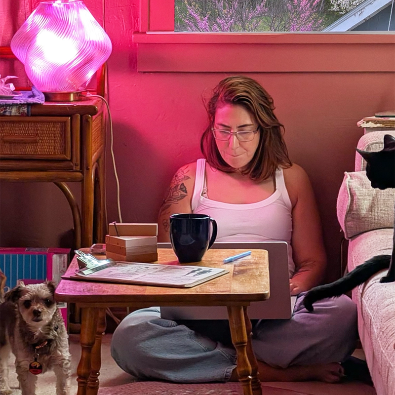 Daniela Bloch sits cross-legged on the floor of a pink room working on a laptop at a small wooden table. A glowing pink lamp, a coffee mug, papers, and a small dog are nearby, with a window overlooking trees behind her.
