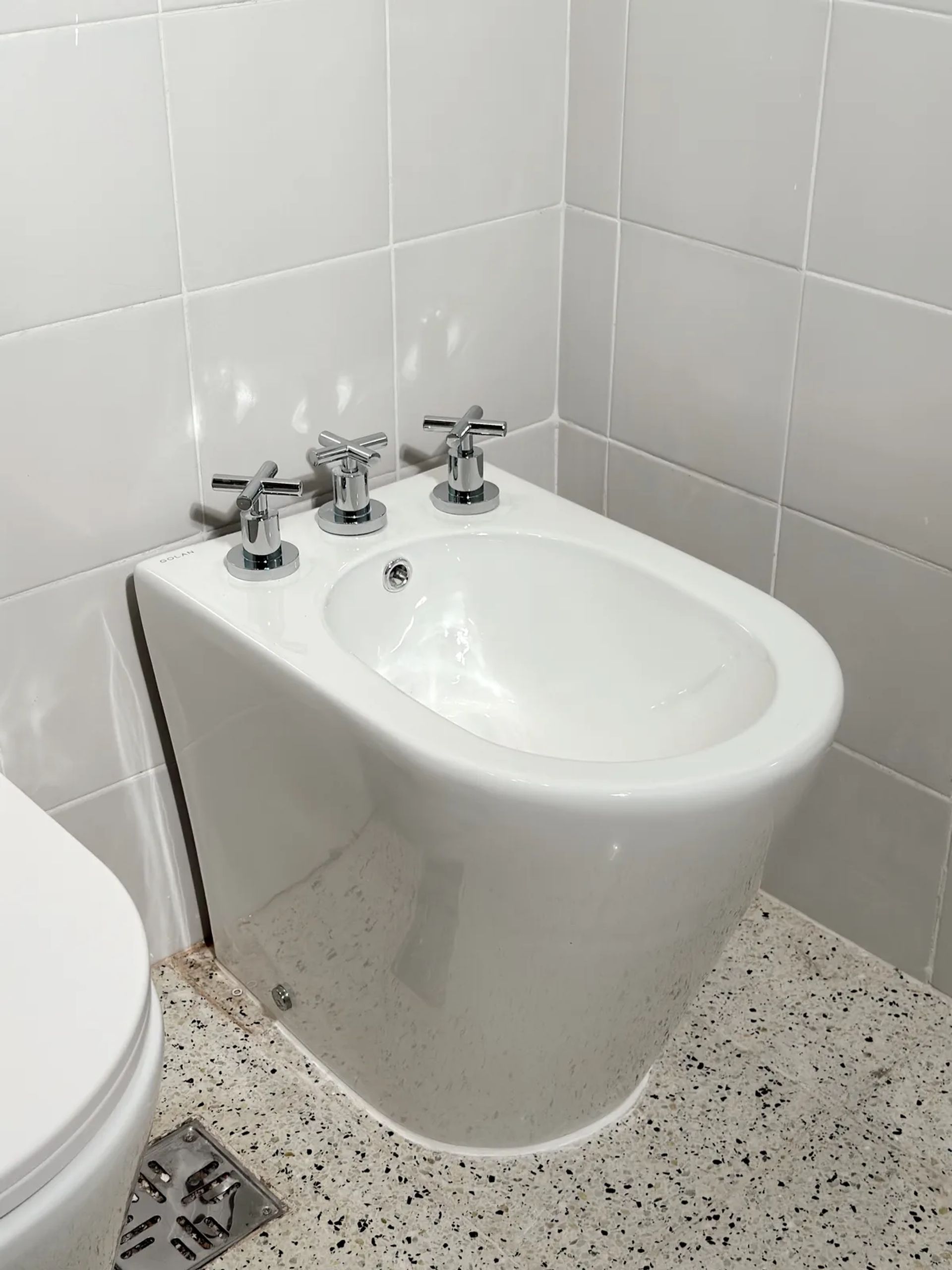 Bidet blanco con grifería cromada de cruz, sobre piso granítico y pared de azulejos blancos