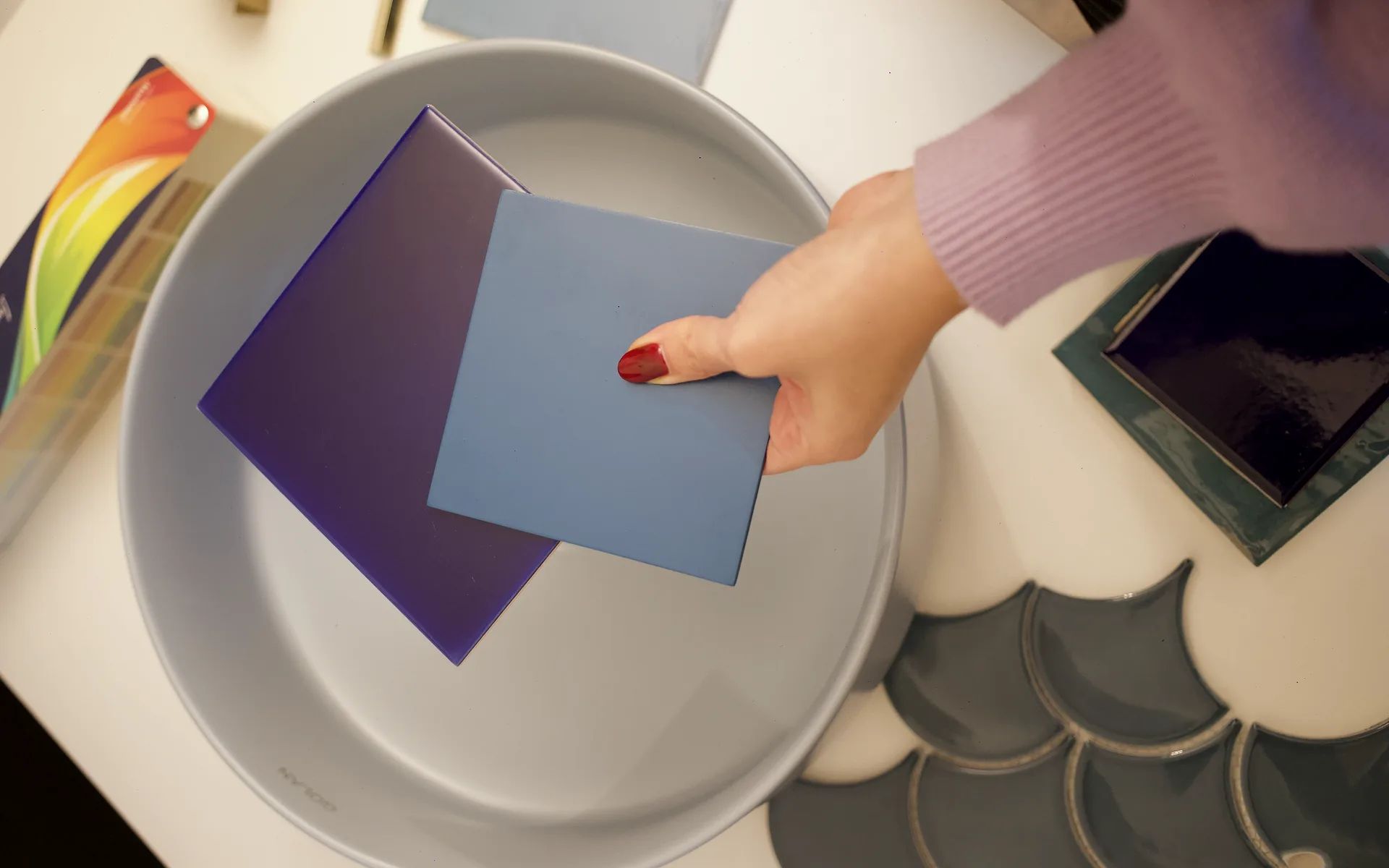 Vista cenital de una bacha gris claro sobre la mesa de materiales, con azulejos violeta y celeste en la mano, cerámicos escama y una carta de colores