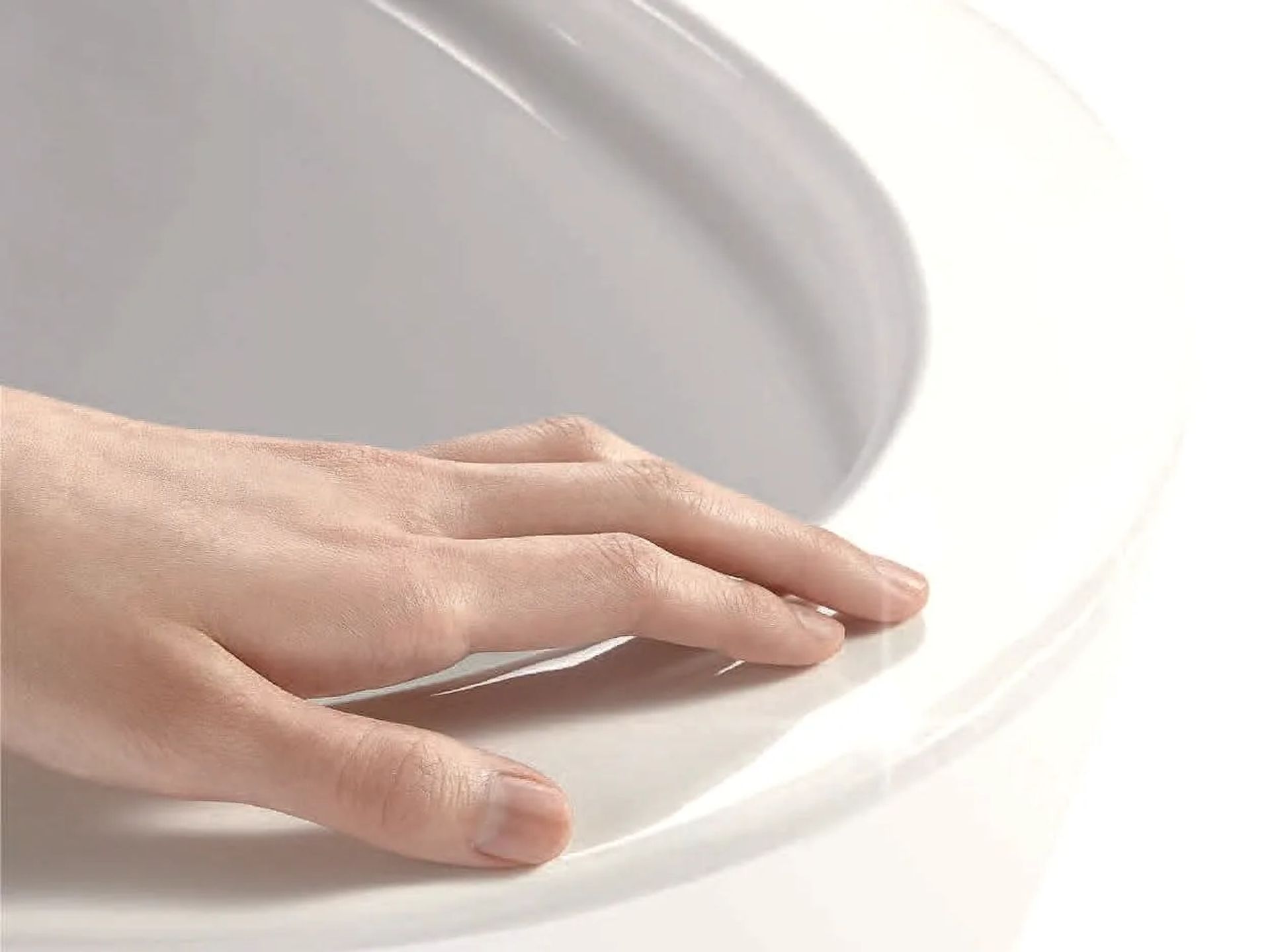 Mano apoyada sobre el borde de un inodoro blanco sin aro interior: el esmalte baja continuo hacia el interior del cuenco