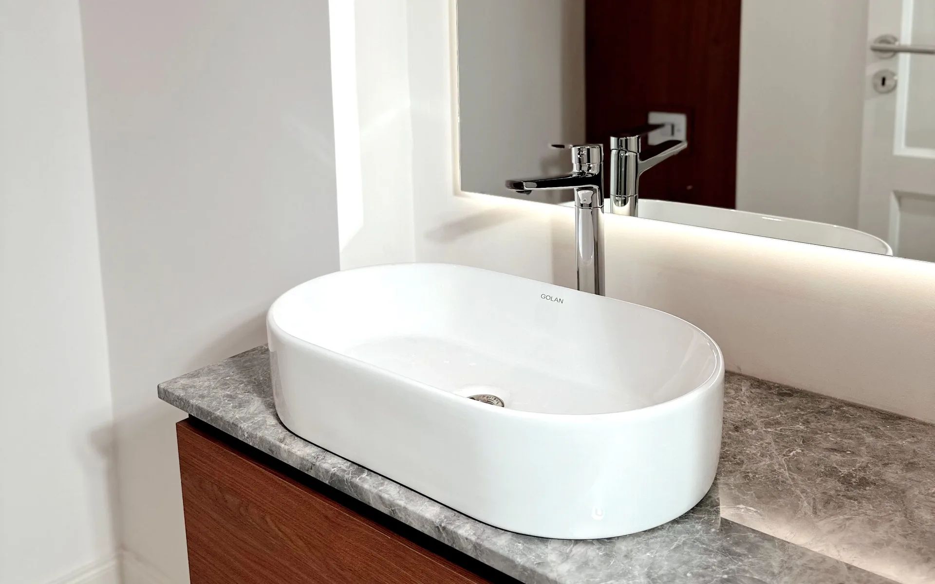 Bacha de apoyo ovalada blanca sobre una mesada de piedra gris, con mueble de madera y espejo iluminado