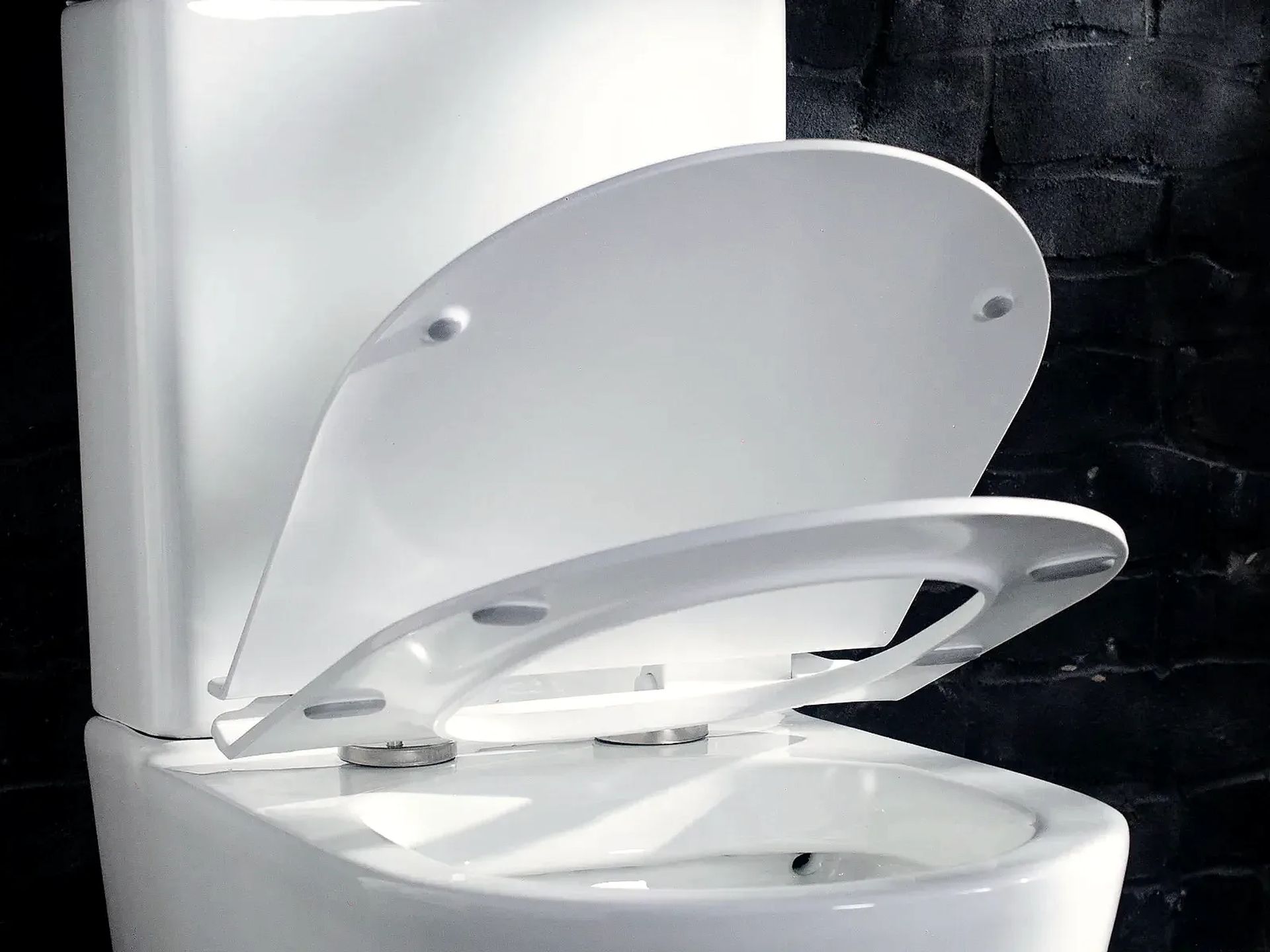 Detalle de la tapa y el asiento de plástico UF levantados sobre un inodoro Symphony blanco, con las bisagras metálicas, frente a una pared de ladrillo negro