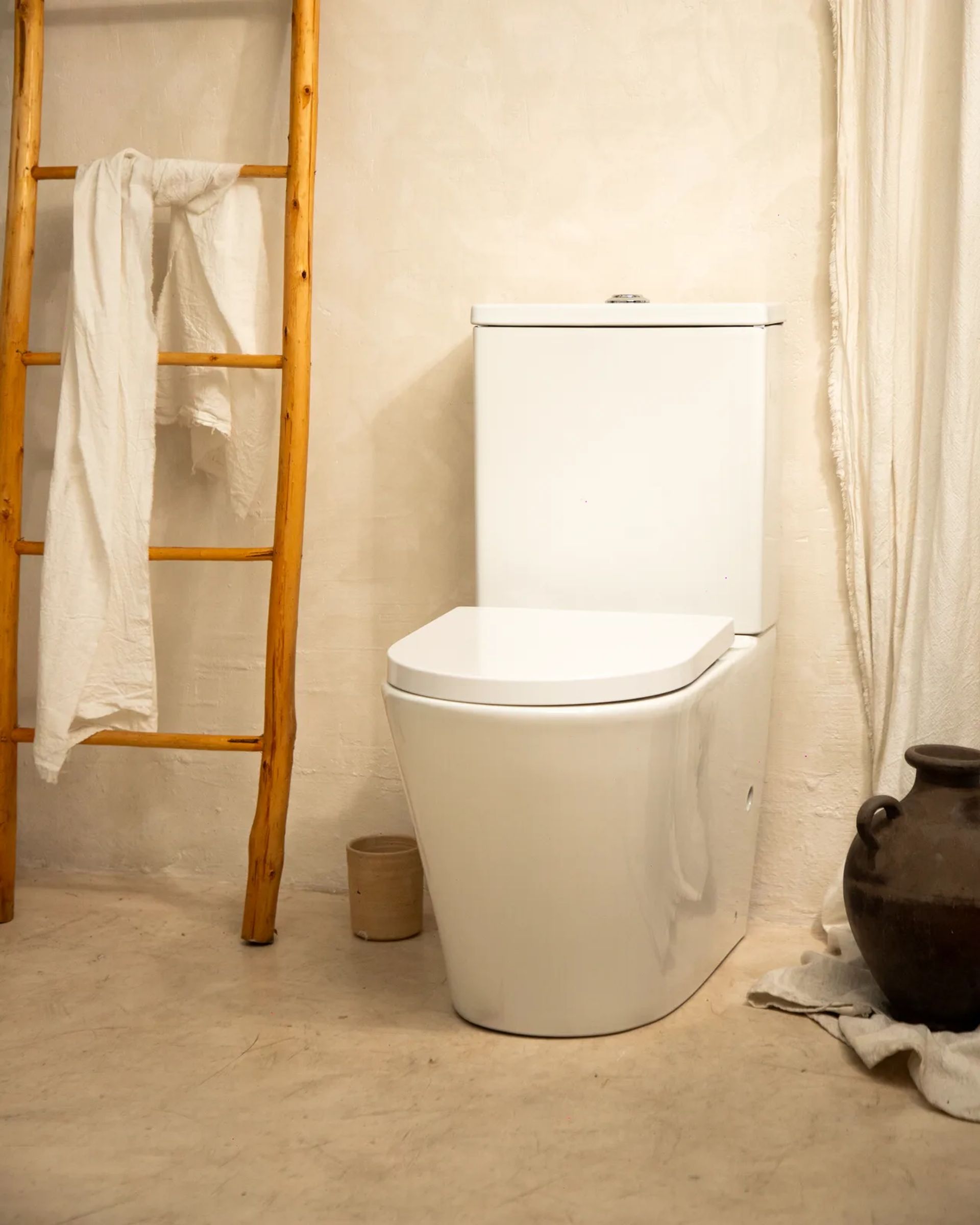 Inodoro ICON blanco en un baño de revoque claro, con una escalera de madera con toallas y una vasija