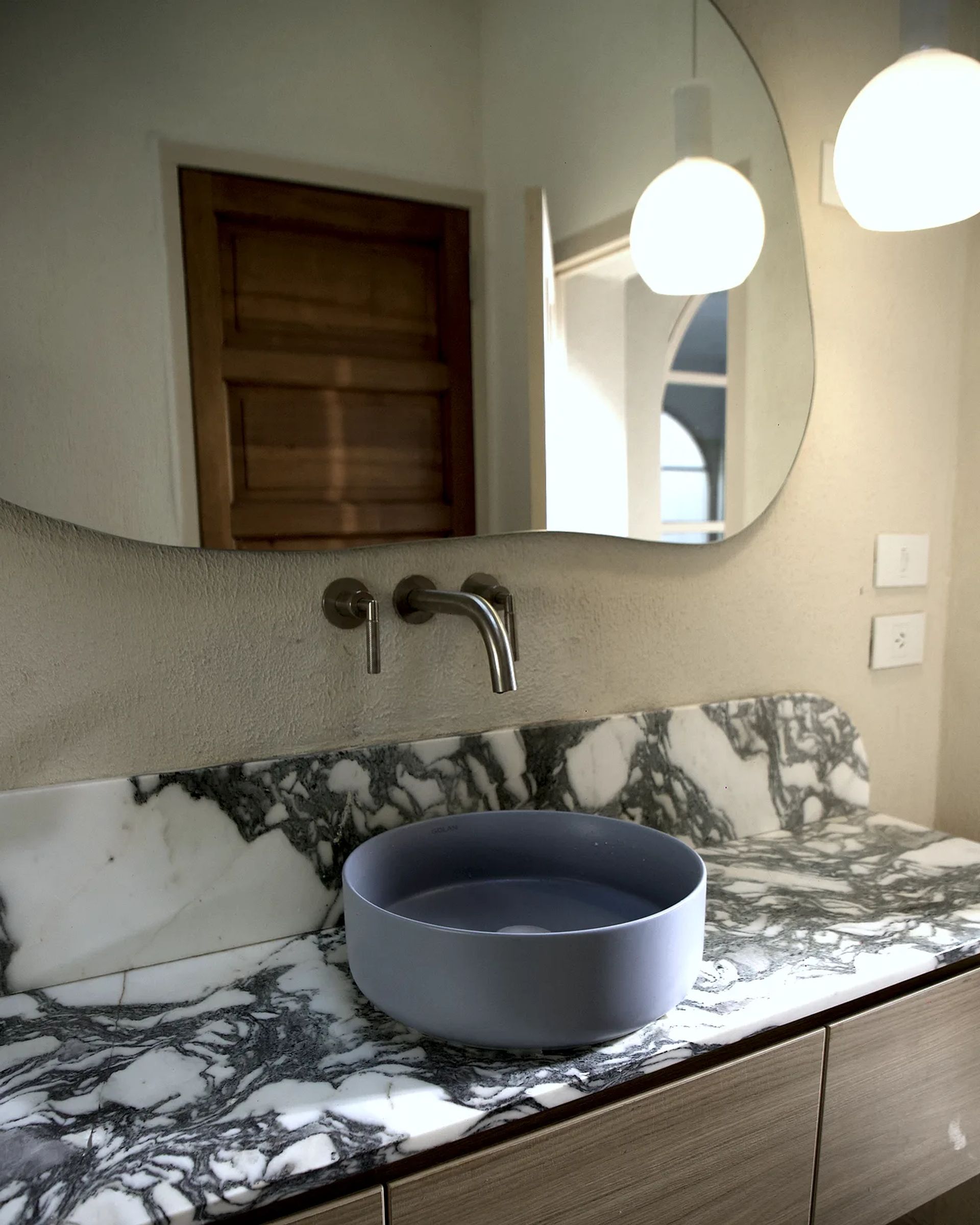 Baño de Casa Lavanda: espejo de forma libre con dos lámparas de globo, grifería de pared y la bacha Picasso celeste sobre la mesada de mármol veteado