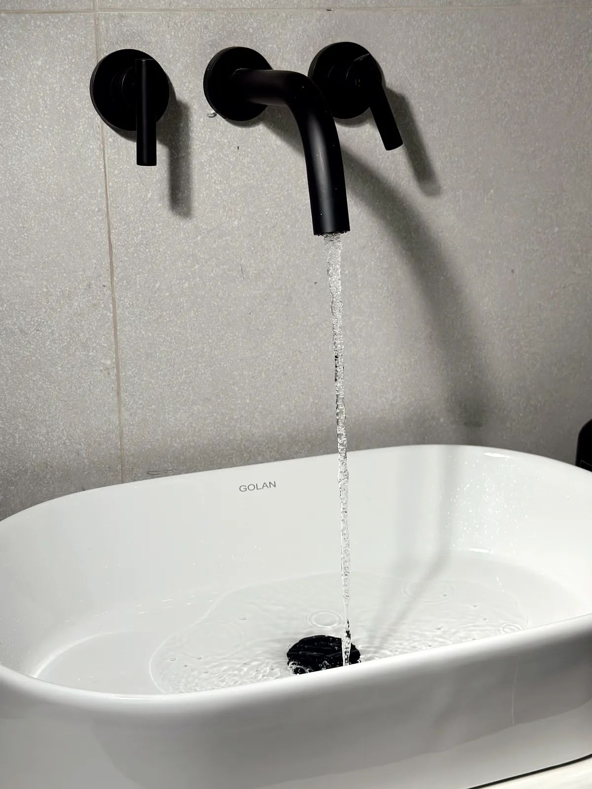 Agua saliendo de la grifería de pared negra sobre la bacha blanca con el logotipo GOLAN
