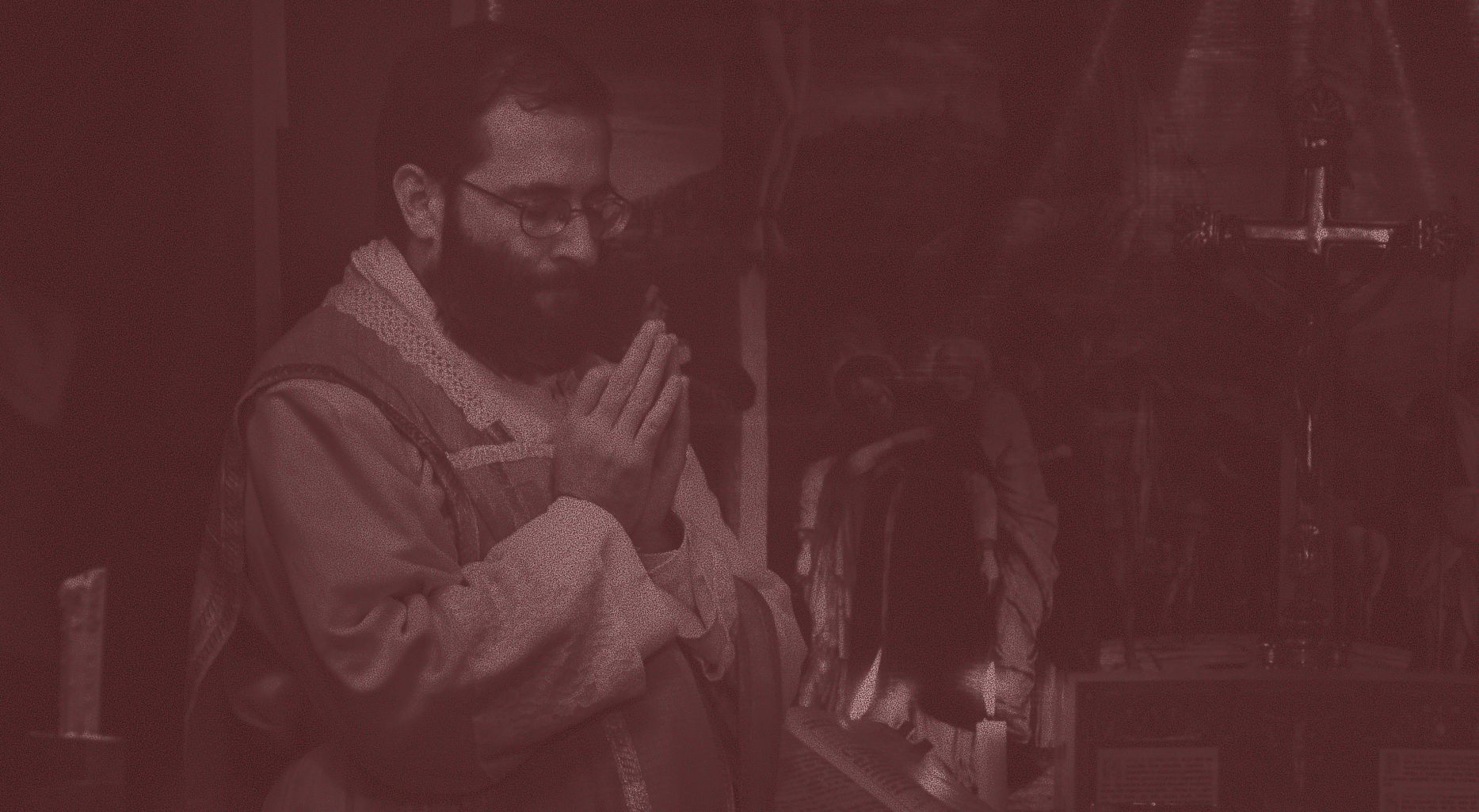 A bearded priest in vestments stands at the altar with his hands joined in prayer, a crucifix and statues behind him.