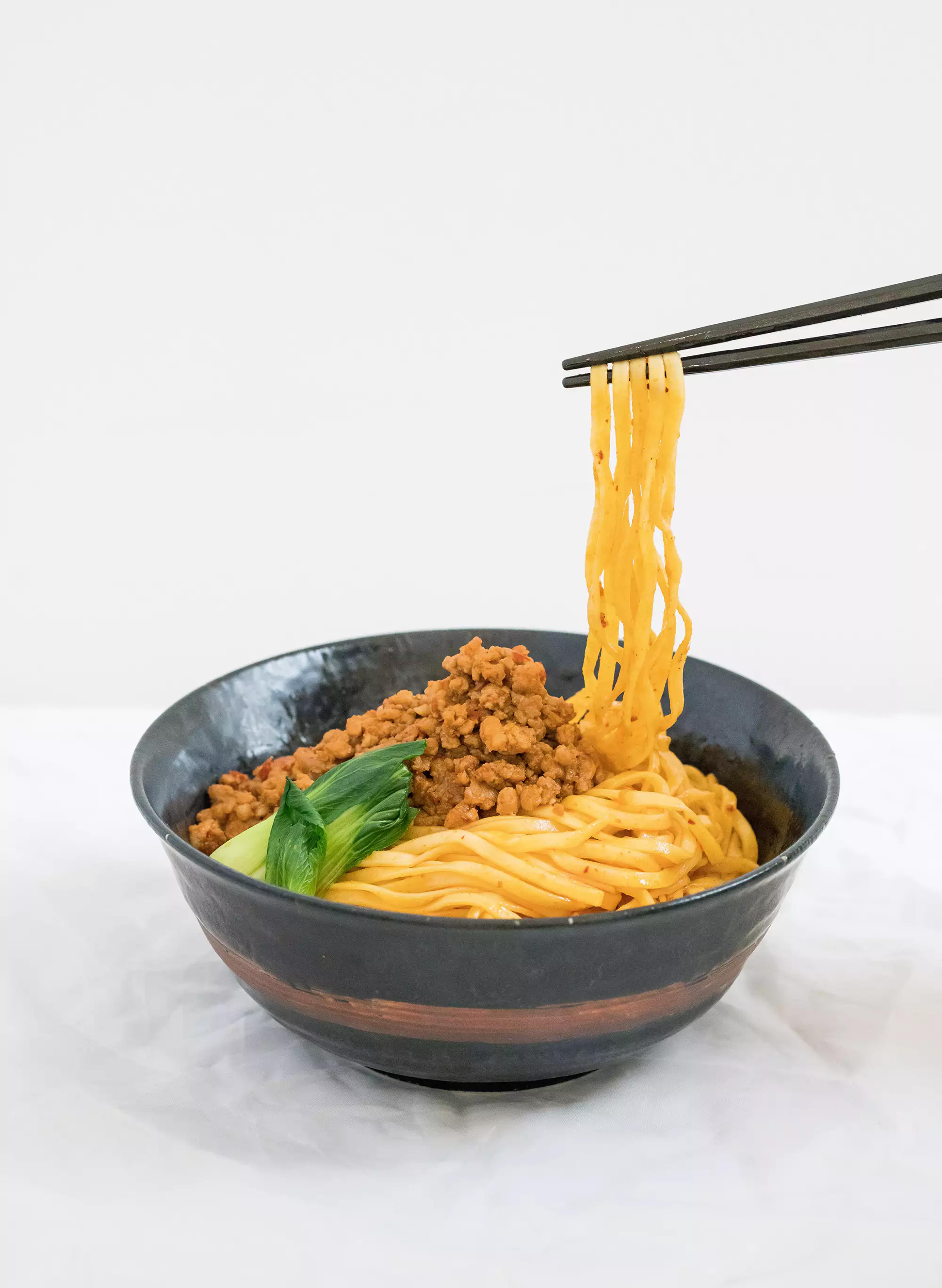 Spicy sesame mazemen on a bowl with protein and bok choi and noodles being pulled with chop sticks.