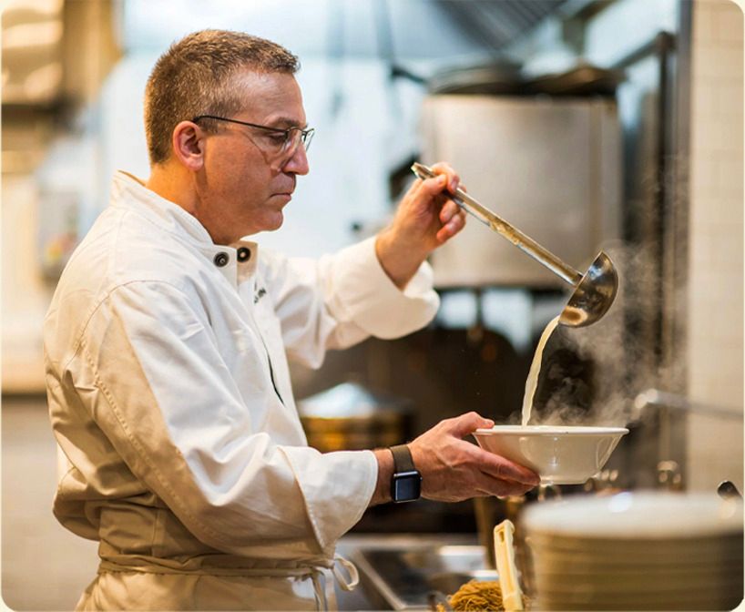 Chef ladling broth into a bowl of ramen in a restaurant kitchen