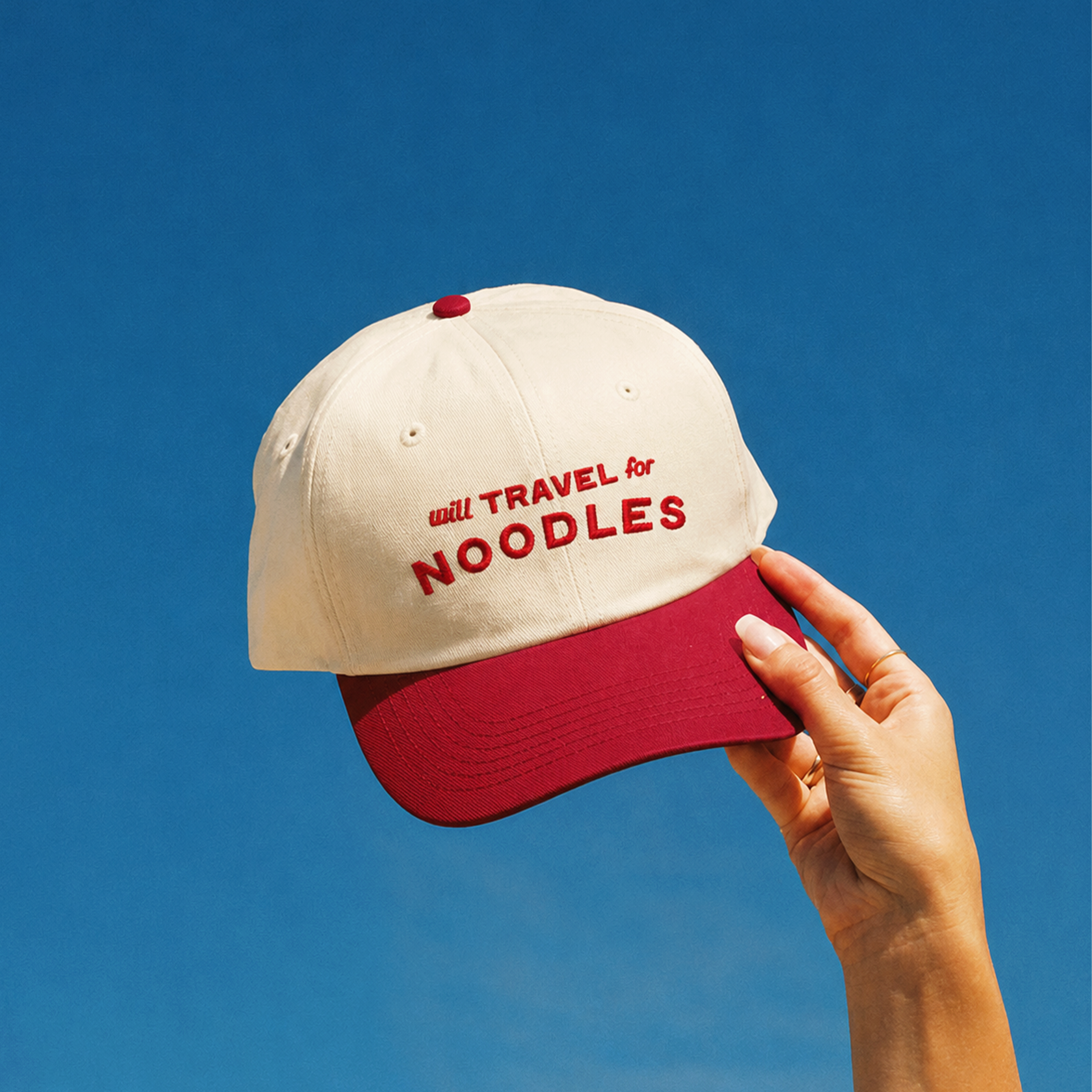 Cream 5-panel snapback cap with a red brim and red top button, embroidered with "will TRAVEL for NOODLES" in red lettering across the front.