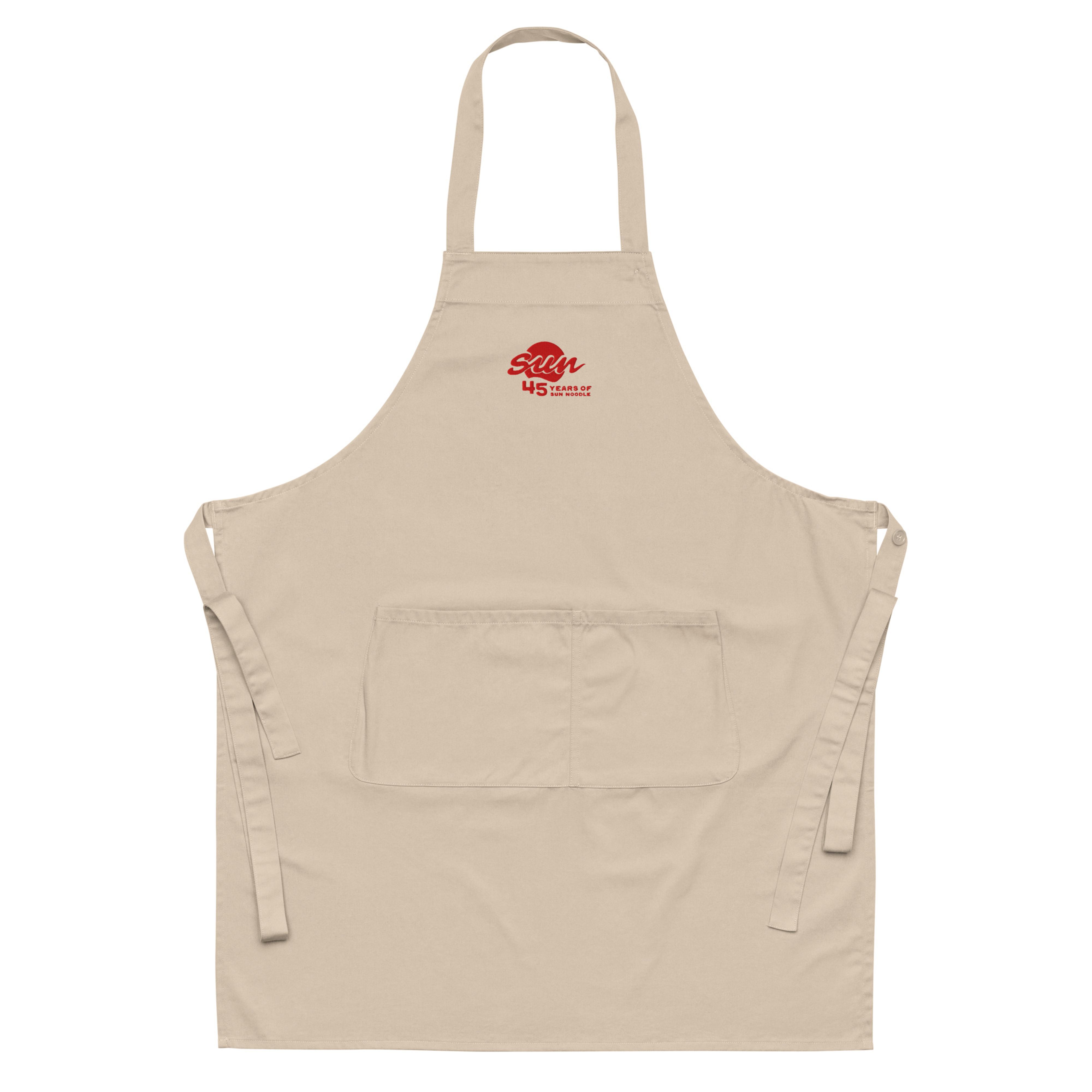 Beige organic cotton apron with a neck loop, waist ties, and a front pocket, embroidered near the chest with the red "45 Years of Sun Noodle" logo featuring a stylized "sun" script over a red circle.