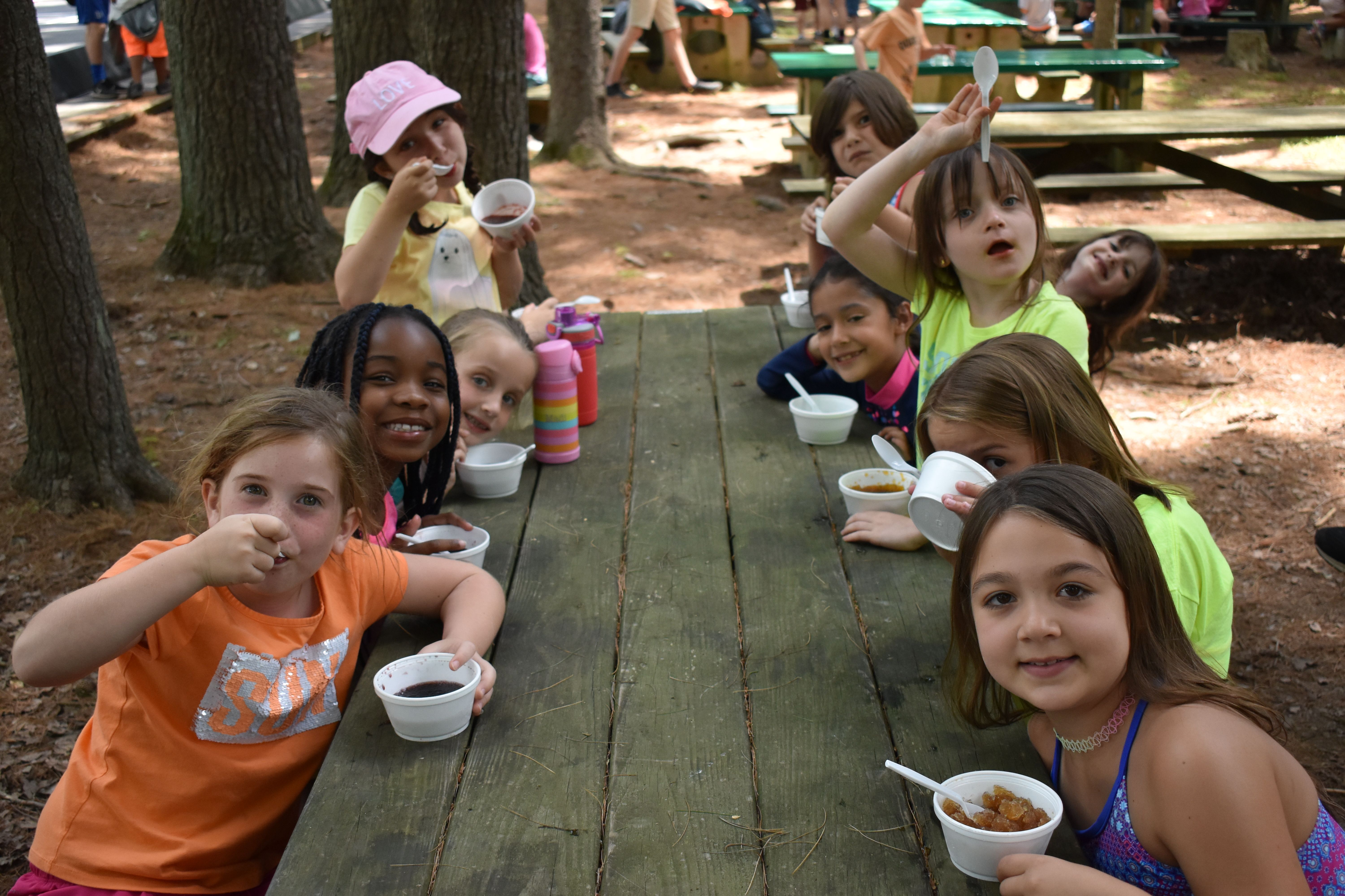 kids sitting around a picnic table eating and smiling at the camera at Rambling Pines Day Camp