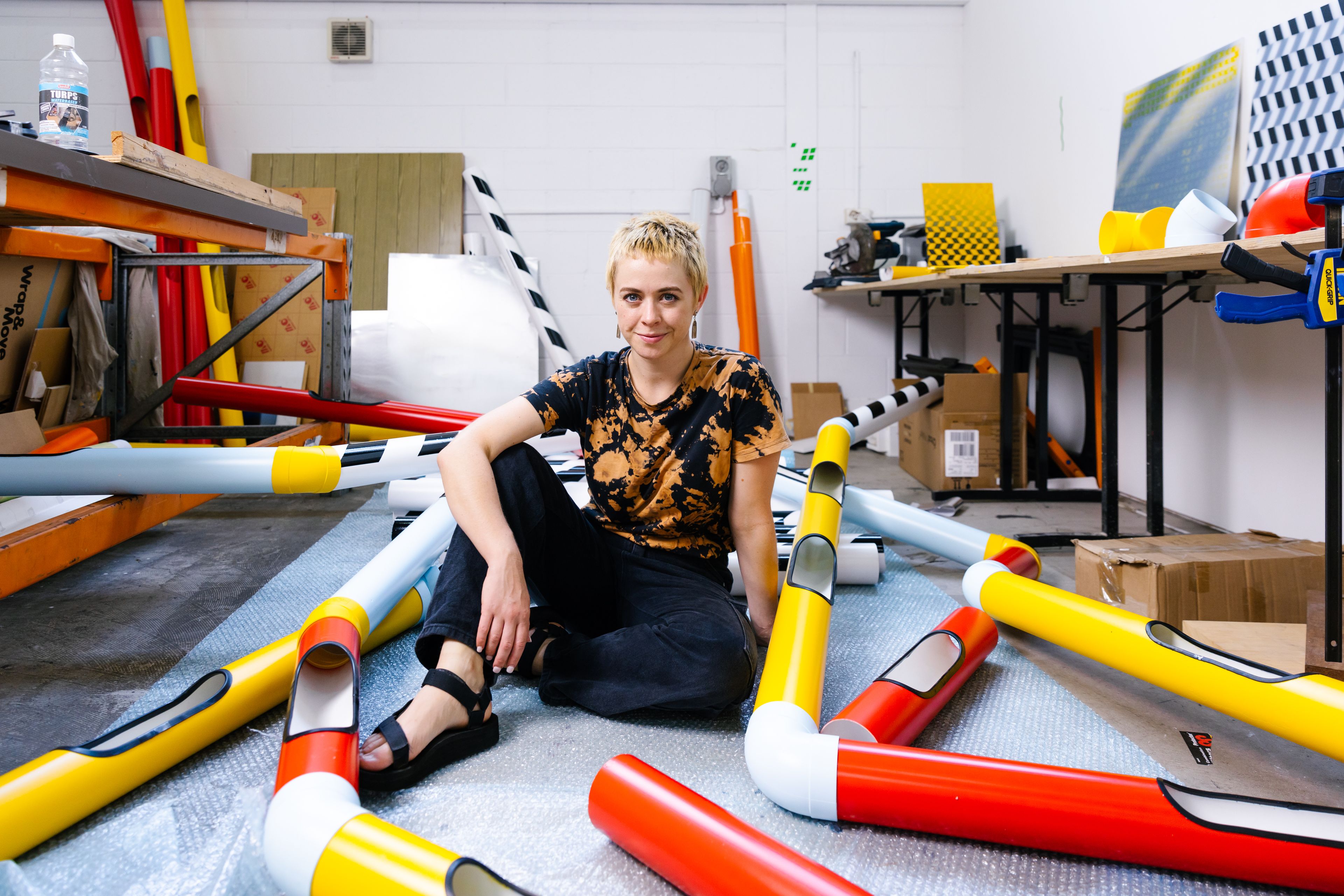 Artist Nadia Odlum in her studio surrounded by bright red and yellow pipes and sculptural objects.
