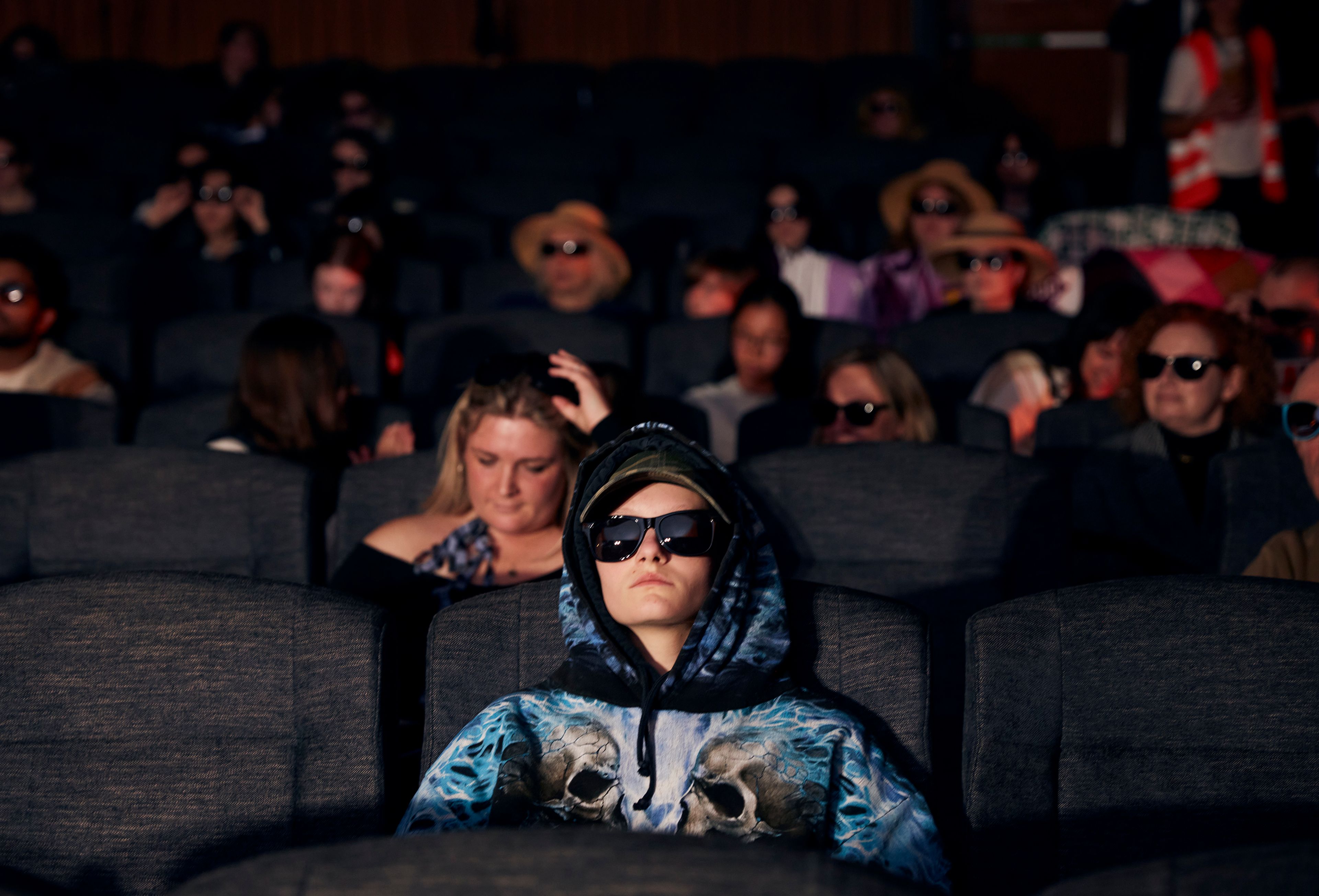 A young man in a hoodie and sunglasses leans back in his cinema seat watching the screen