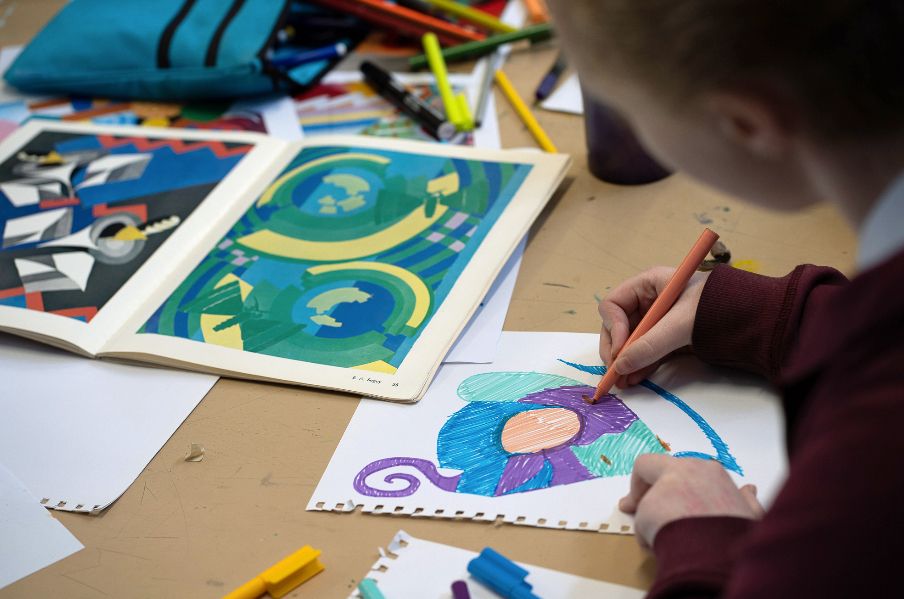 A teenager drawing a colourful abstract picture, on a cluttered desk covered in reference images and marker pens.