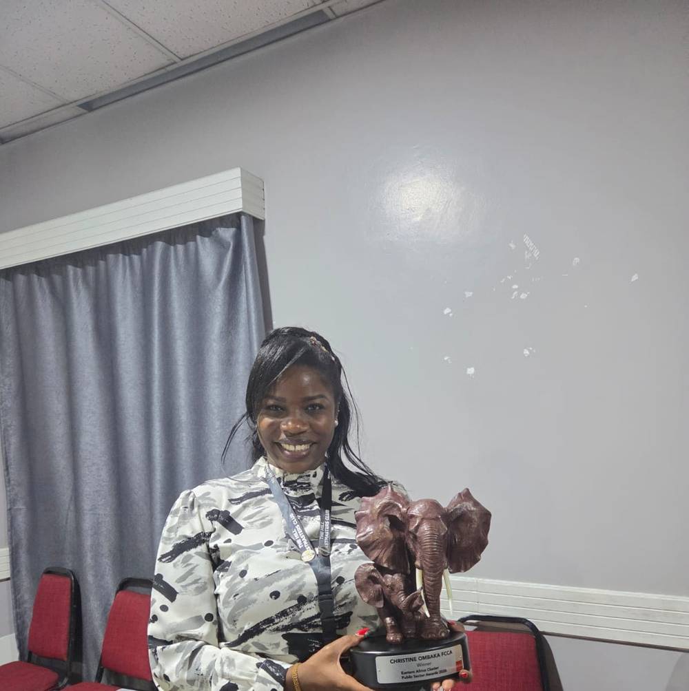 Christine Ombaka in the club meeting room, smiling and holding the elephant trophy, with rows of red chairs behind her.