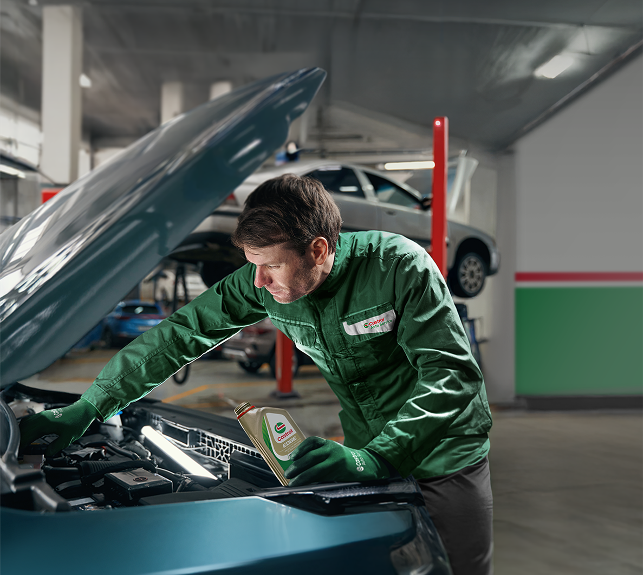 Mekaniker i Castrol Service-jakke der tjekker under motorhjelmen på en bil, mens han holder en flaske Castrol Edge-olie