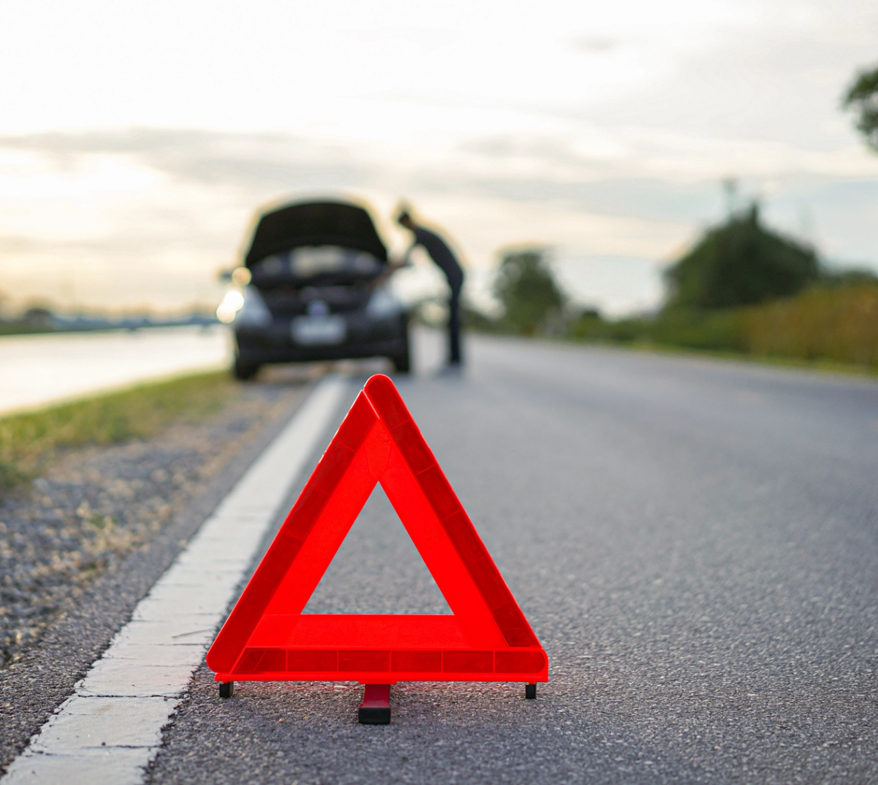 Rødt advarselstrekant ved vejkanten foran en bil med åben motorhjelm, hvor en person tjekker motoren.
